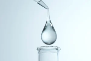 A glass dropper releases a large droplet into a narrow-neck vial in a lab setting, with pale blue lighting above.