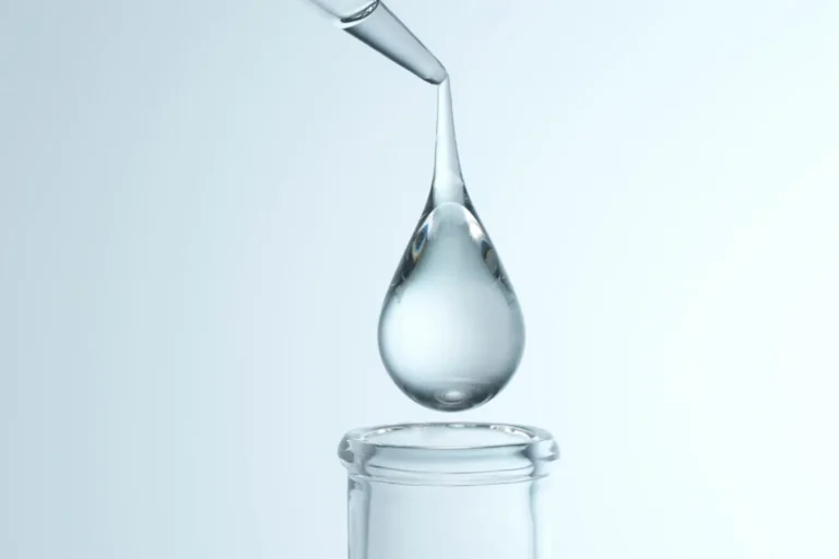 A glass dropper releases a large droplet into a narrow-neck vial in a lab setting, with pale blue lighting above.