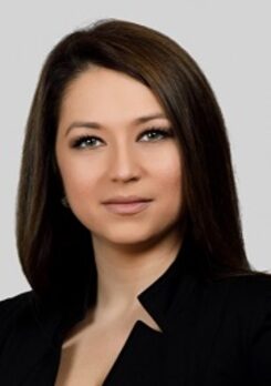 1709137384 Professional headshot of a woman with dark brown hair in a black blazer in front of a gray background.