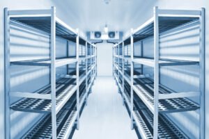 Aisle of empty stainless steel shelving units in a walk-in refrigerated storage room.