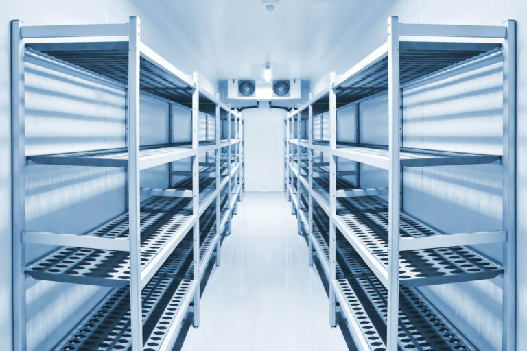 Aisle of empty stainless steel shelving units in a walk-in refrigerated storage room.
