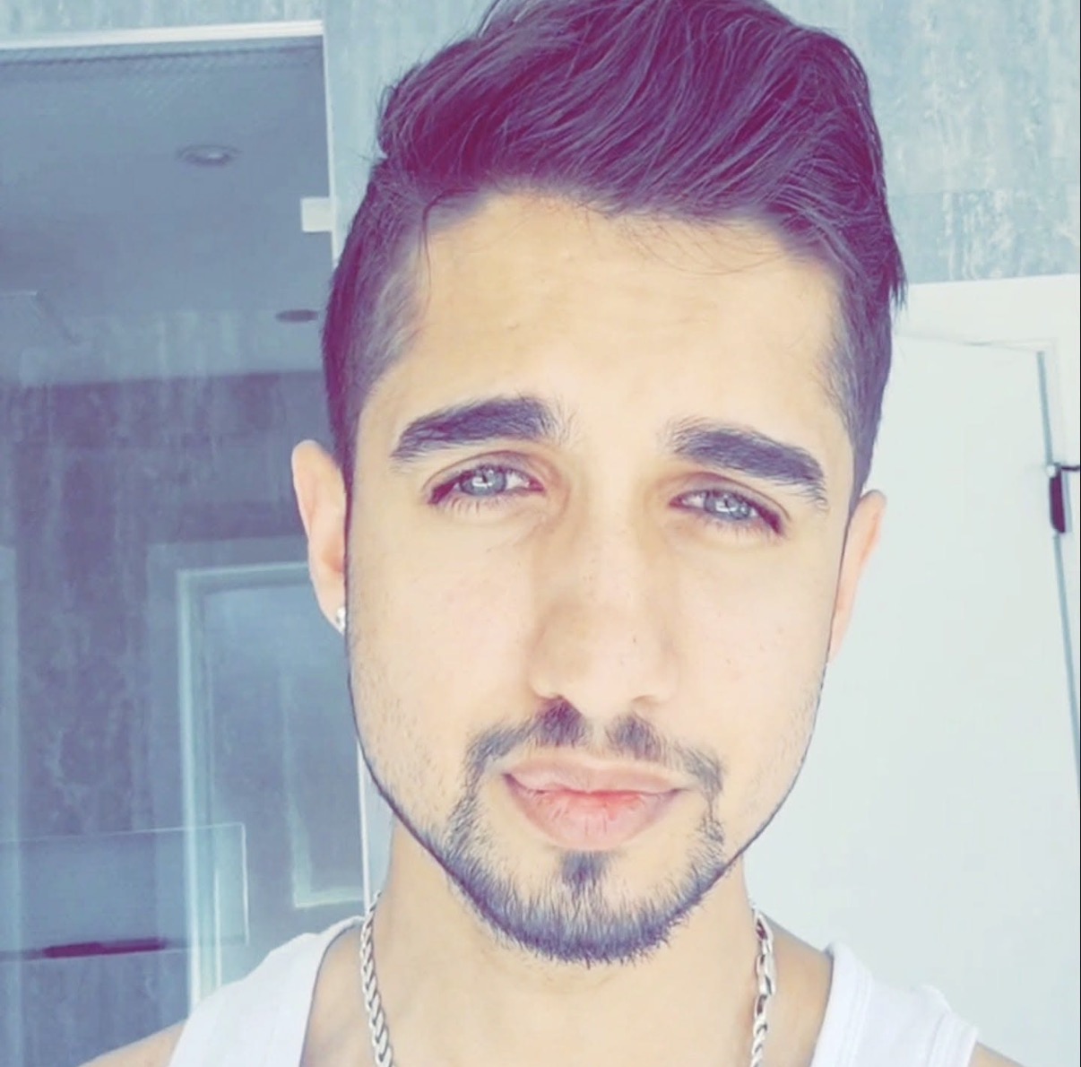 Screenshot Close-up portrait of a young man with blue eyes, dark styled hair, light stubble, wearing a white tank top.
