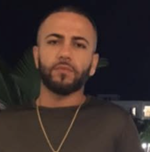 Screenshot 2026-09-27 at 23341 PM Young man with a trimmed beard and short hair wearing a dark shirt and gold chain, photographed at night with a dark background.