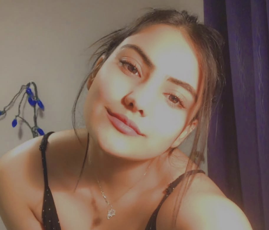 Screenshot 2026-09-27 at 24506 PM Young woman taking a selfie indoors, wearing a black tank top and a silver necklace, with a soft smile and purple curtains in the background.