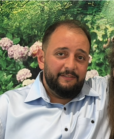 View recent photos 2 Close-up of a man with short dark hair and a beard, wearing a light blue button-down shirt, standing in front of blooming flowers and greenery.