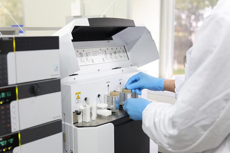Lab technician in blue gloves loading sample tubes into a benchtop analytical instrument in a clean lab environment.